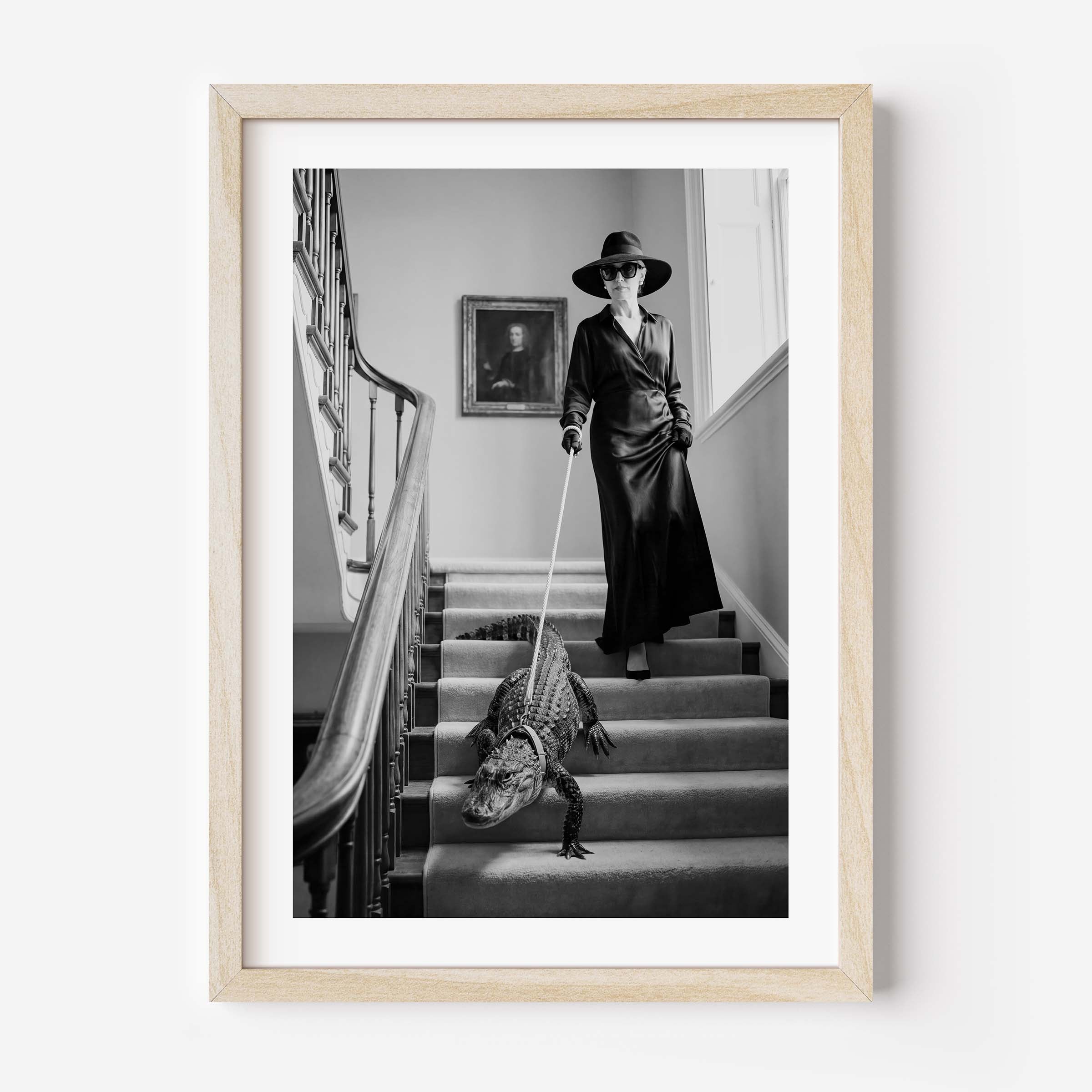 A framed black and white art print depicting a woman in a long coat and hat walking a crocodile on a staircase, enclosed in a light wood frame against a white wall.