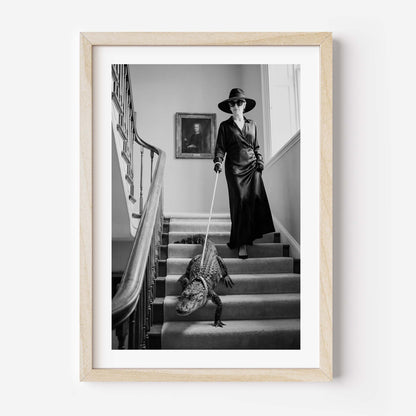 A framed black and white art print depicting a woman in a long coat and hat walking a crocodile on a staircase, enclosed in a light wood frame against a white wall.