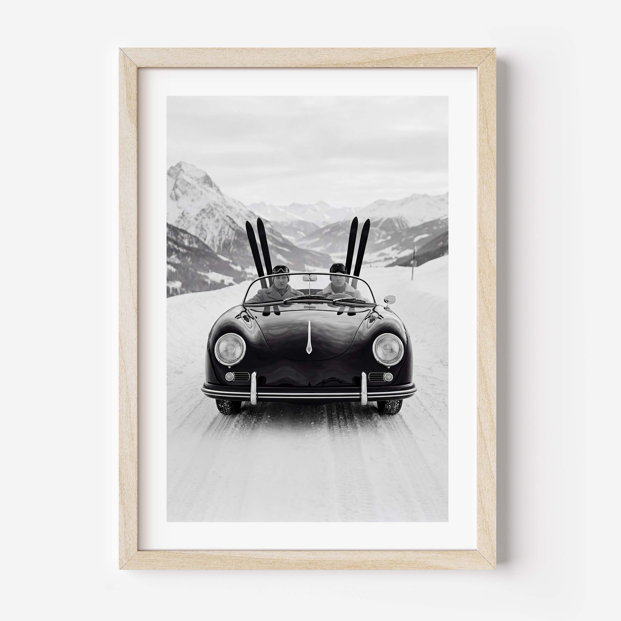 Black-and-white photo of a vintage convertible driving on a snowy mountain road, with two people inside and skis standing upright behind them, alpine peaks in the background.