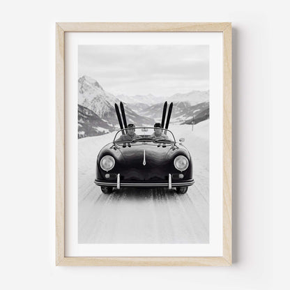 Black-and-white photo of a vintage convertible driving on a snowy mountain road, with two people inside and skis standing upright behind them, alpine peaks in the background.