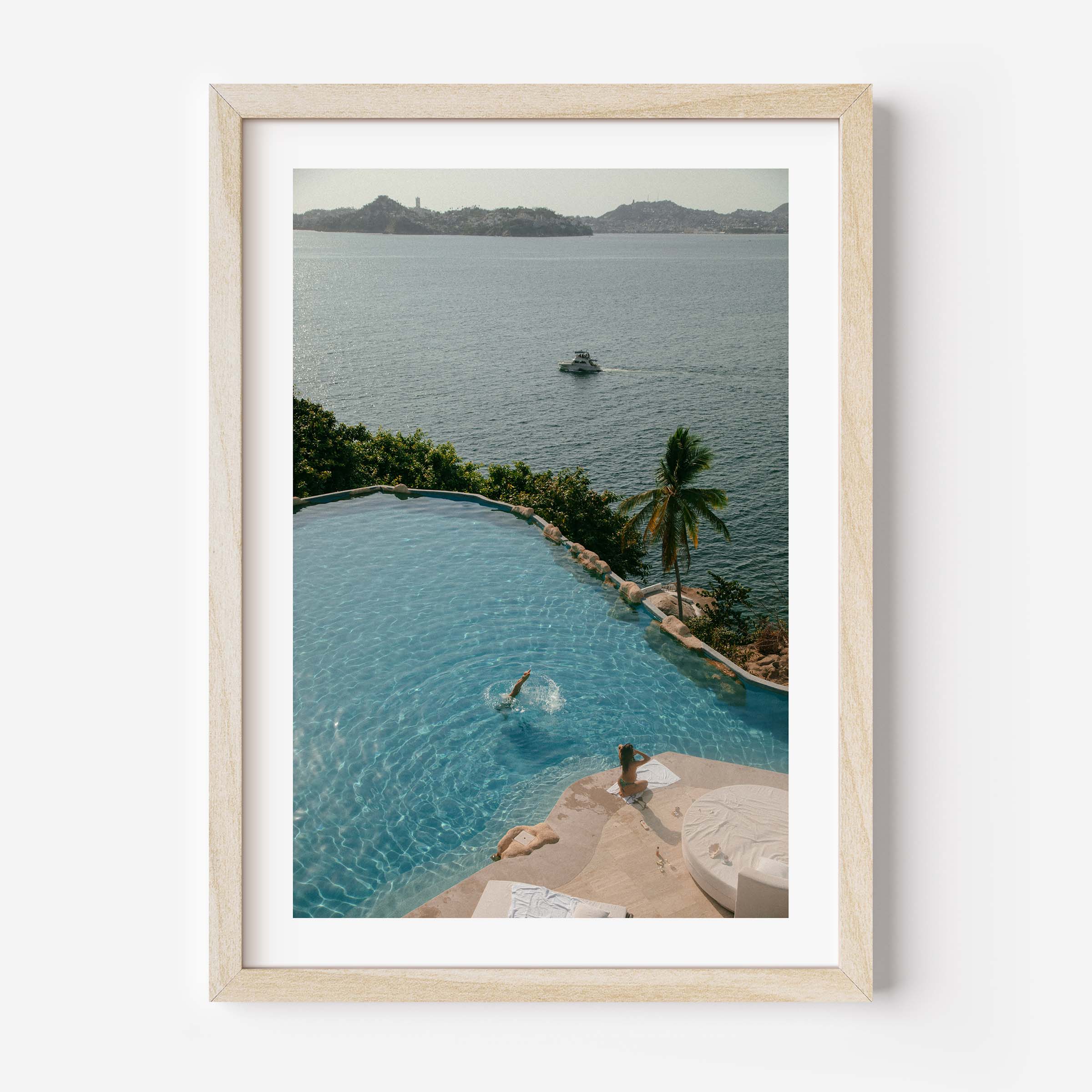 Framed color photograph of a diver in a turquoise infinity pool above a tropical bay with palm tree, sunlounger and boat in the distance — by Britt Van der Meyden.
