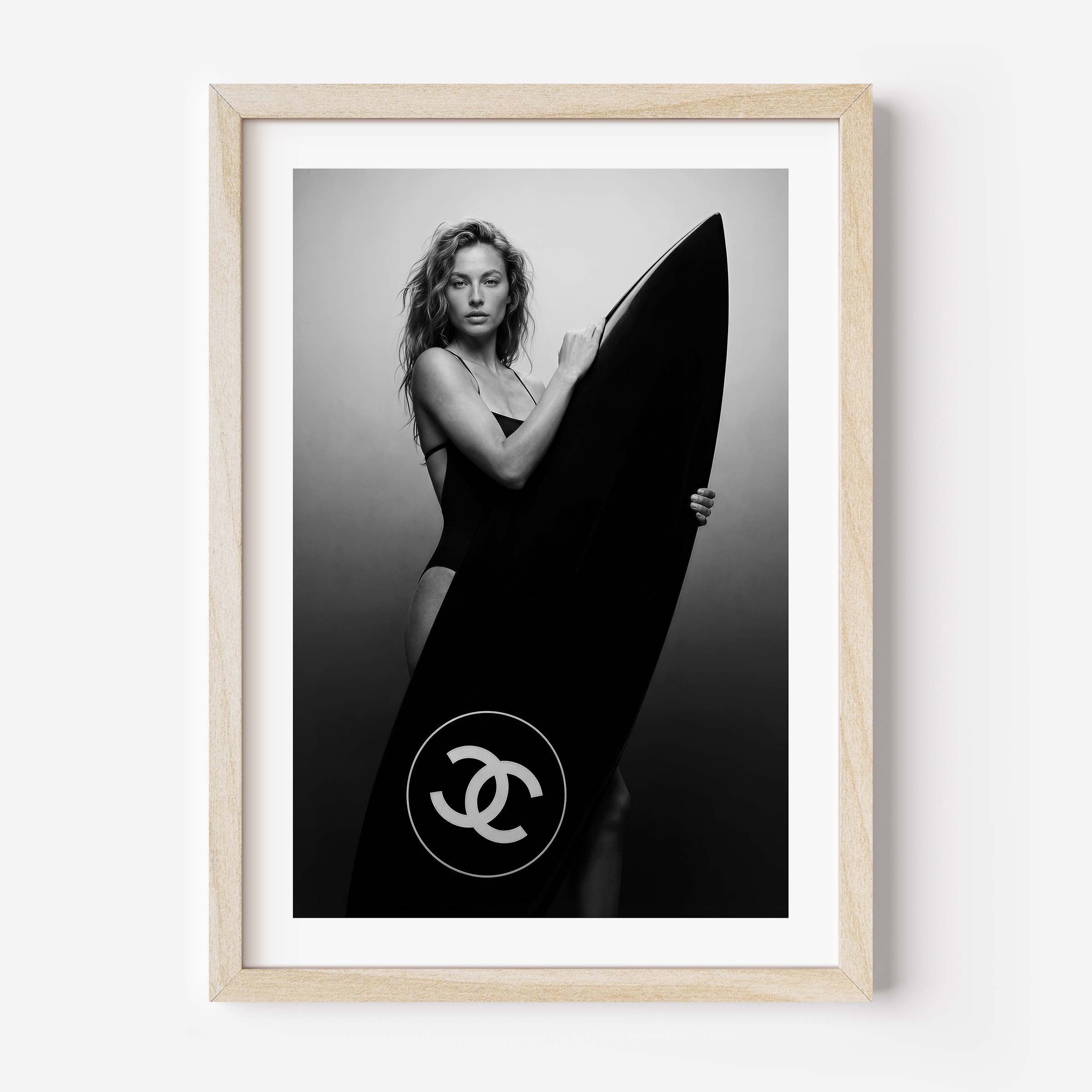 Black-and-white fine art photo of a woman in a swimsuit holding a large surfboard featuring a circular double-C style logo, against a minimal studio background.
