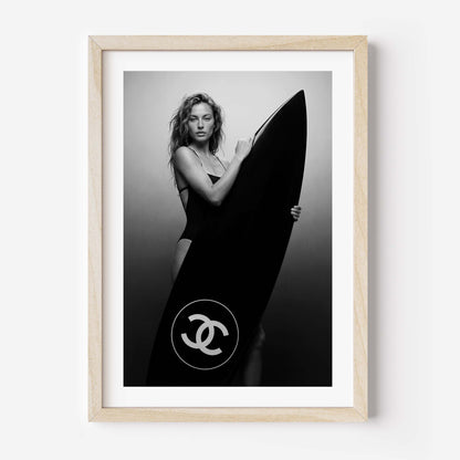 Black-and-white fine art photo of a woman in a swimsuit holding a large surfboard featuring a circular double-C style logo, against a minimal studio background.