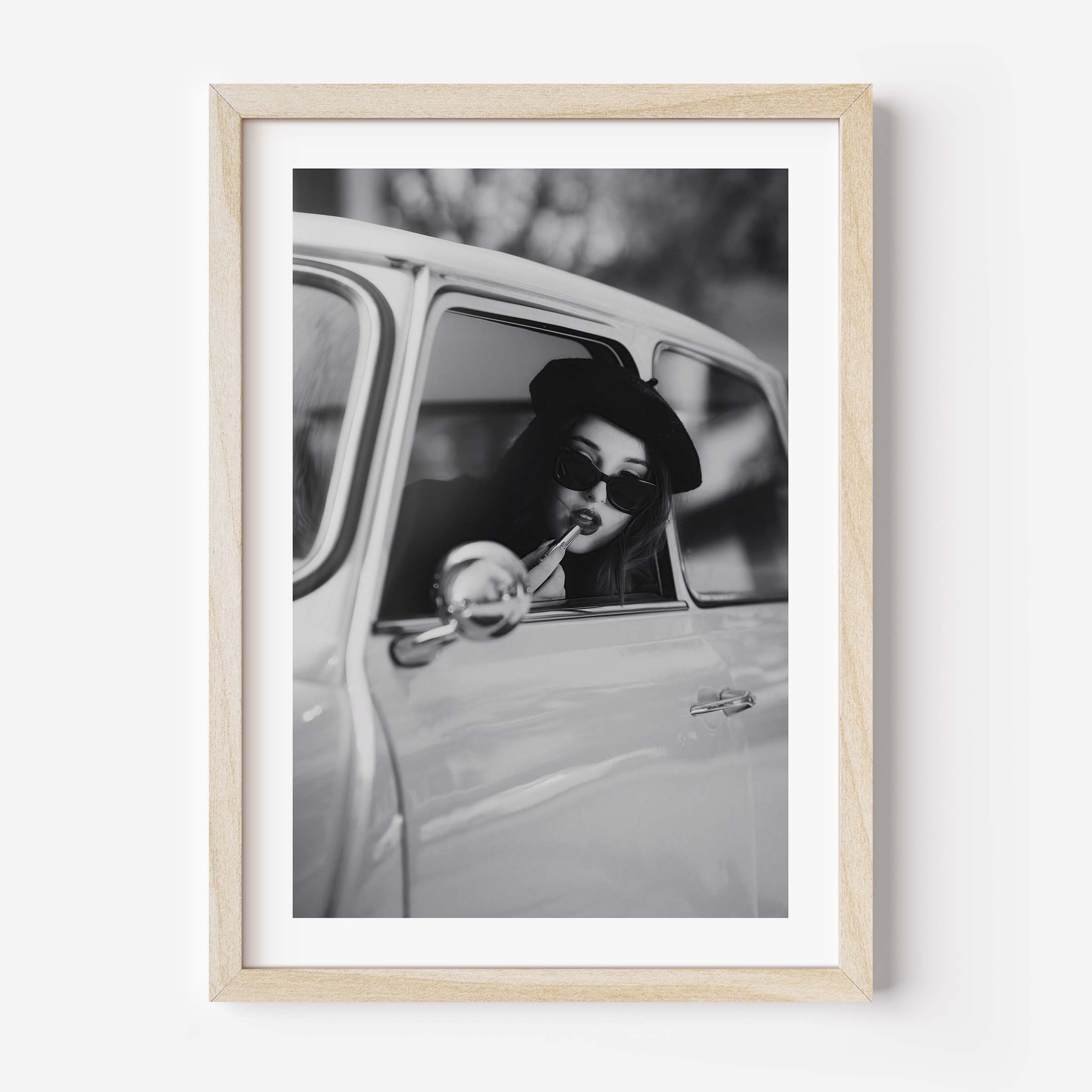 Black and white framed wall art print showing a woman in sunglasses sitting in a vintage car while applying lipstick.