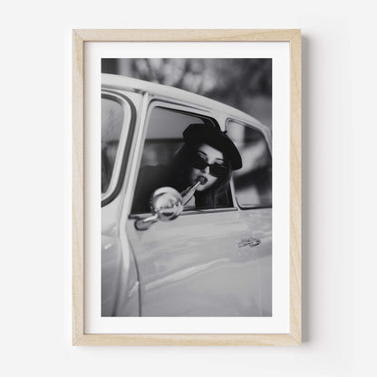 Black and white framed wall art print showing a woman in sunglasses sitting in a vintage car while applying lipstick.