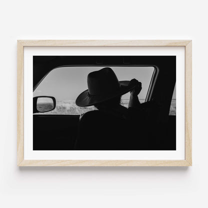 Black-and-white fine art print of a person wearing a cowboy hat seen from inside a car, looking out over an open landscape.
