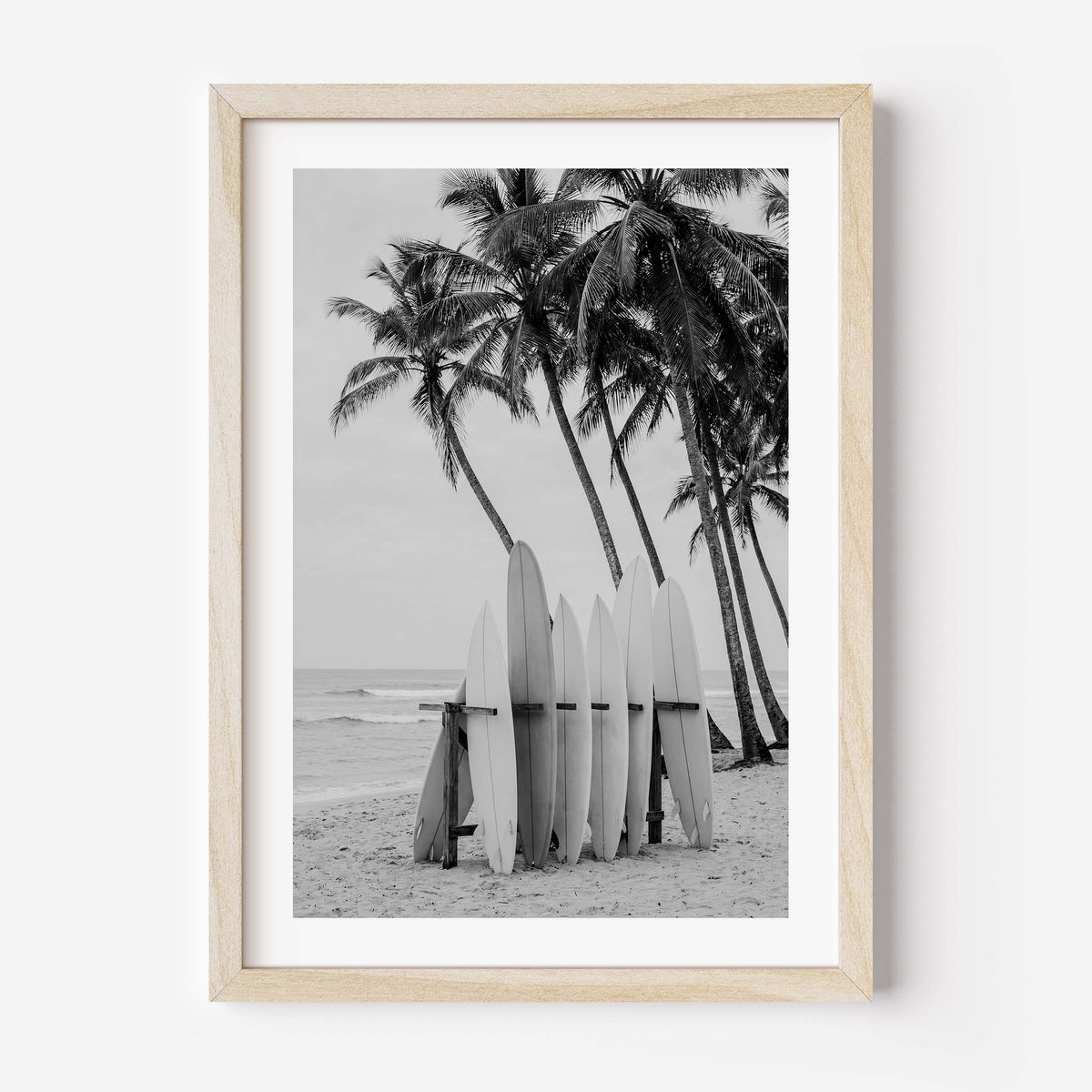 Framed black and white beach photograph featuring palm trees, surfboards, and ocean waves, displayed in a light natural wood frame against a white wall.