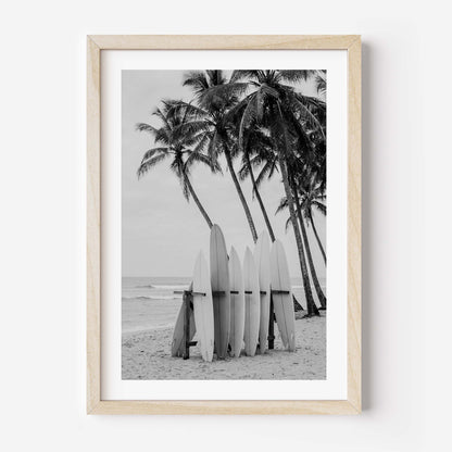 Framed black and white beach photograph featuring palm trees, surfboards, and ocean waves, displayed in a light natural wood frame against a white wall.
