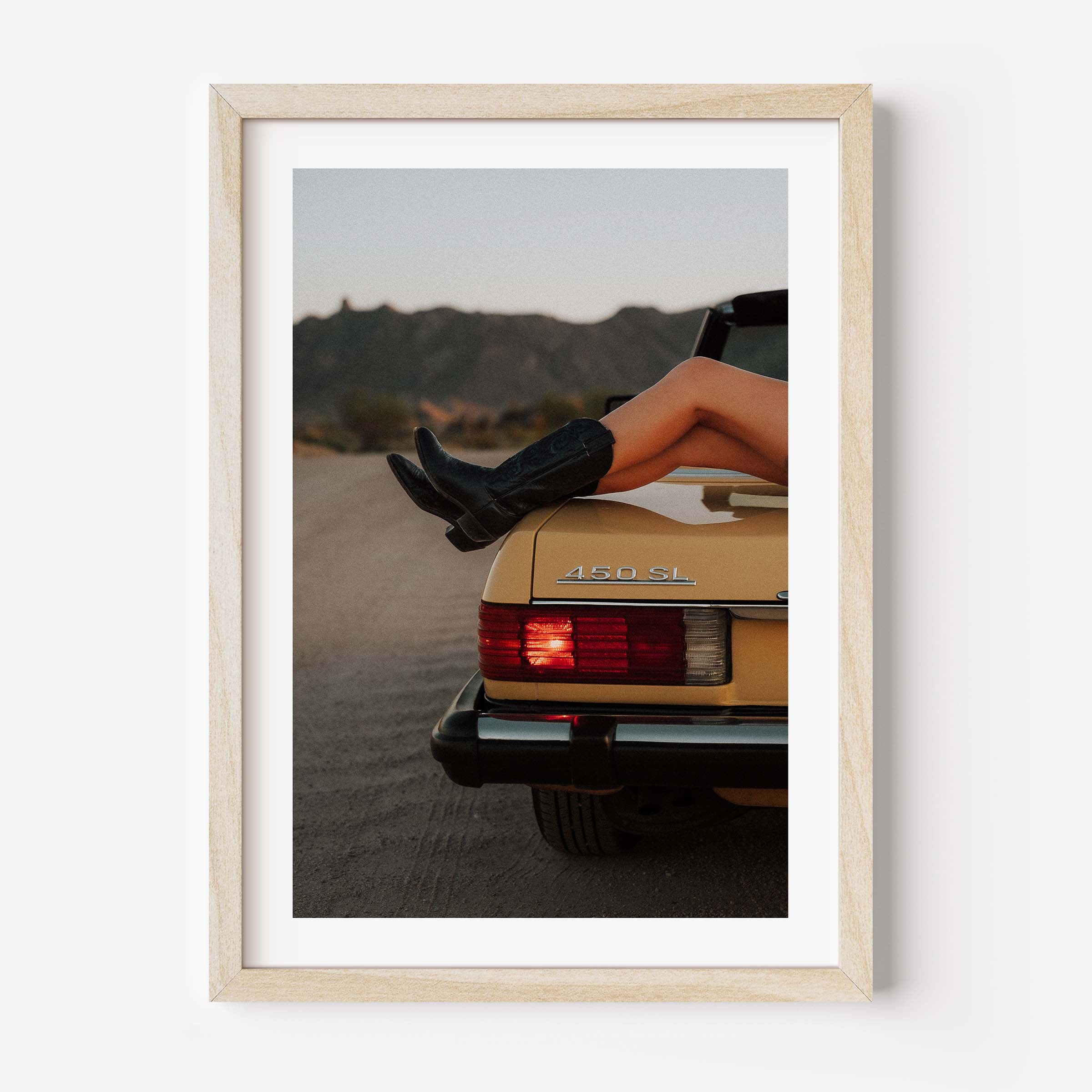 Fine art photo of the rear of a classic Mercedes 450 SL at dusk, with a person’s legs in black boots resting on the trunk and mountains in the background.