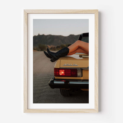 Fine art photo of the rear of a classic Mercedes 450 SL at dusk, with a person’s legs in black boots resting on the trunk and mountains in the background.