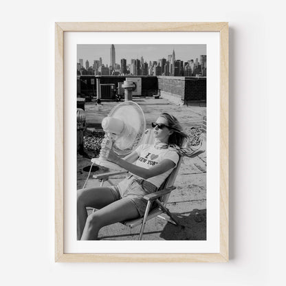 A framed black and white art print featuring a woman sitting on a rooftop chair with a fan, holding a glass of water, with a city skyline in the background. The print is housed in a light natural wood frame with a white mat, shown against a plain white wall.