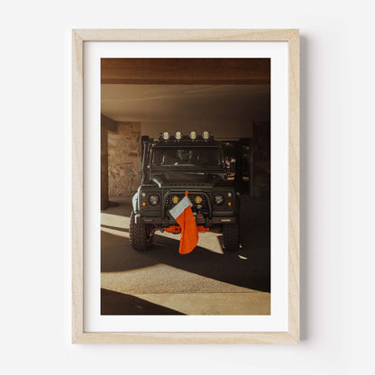 Fine art photo of a Land Rover Defender parked in a garage with warm sunlight, featuring a red Christmas stocking hanging from the front bumper.