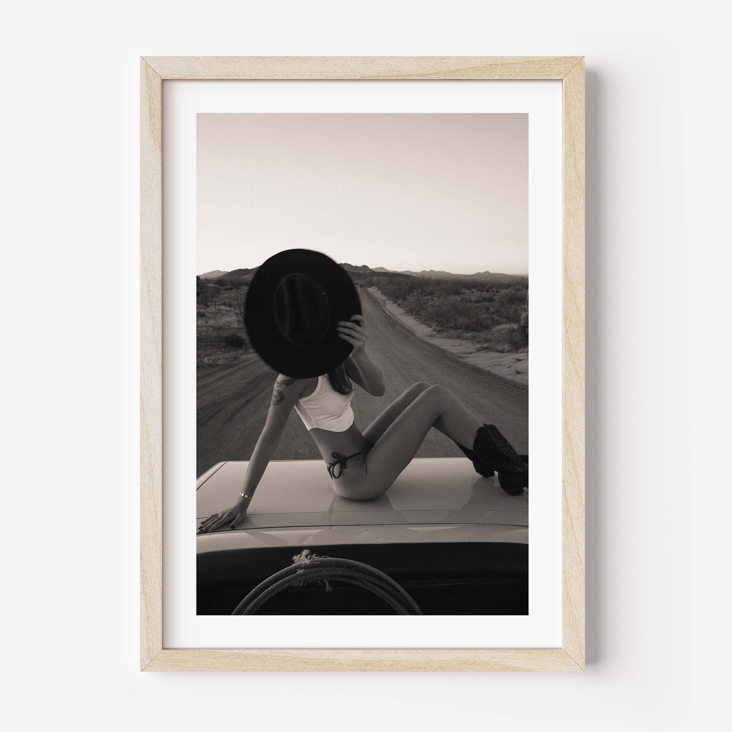 Fine art photo print of a woman sitting on a car roof on a desert road, holding a wide-brim hat in front of her face at sunset.