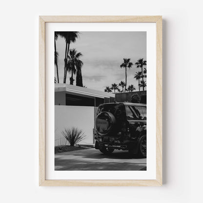 Black-and-white photo of a Land Rover Defender parked in a driveway beside a modern home, with tall palm trees and a bright sky in the background.