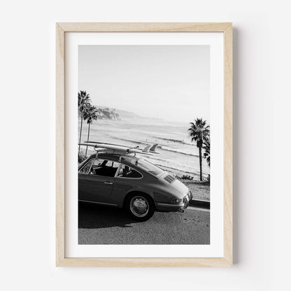 Black and white framed wall art print showing a classic porsche car with surfboards parked by the ocean with palm trees and coastline in the background.