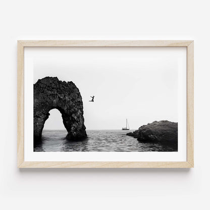 Black and white framed wall art print showing a person jumping into the ocean near a large rock arch with a sailboat in the distance.