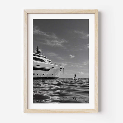 A framed black and white art print depicting a luxury yacht on calm water, with a small diver or swimmer in the distance, presented in a light natural wood frame against a white wall.