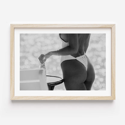 Black-and-white photo of a woman in a white one-piece holding a glass bottle by a sunlit seaside table