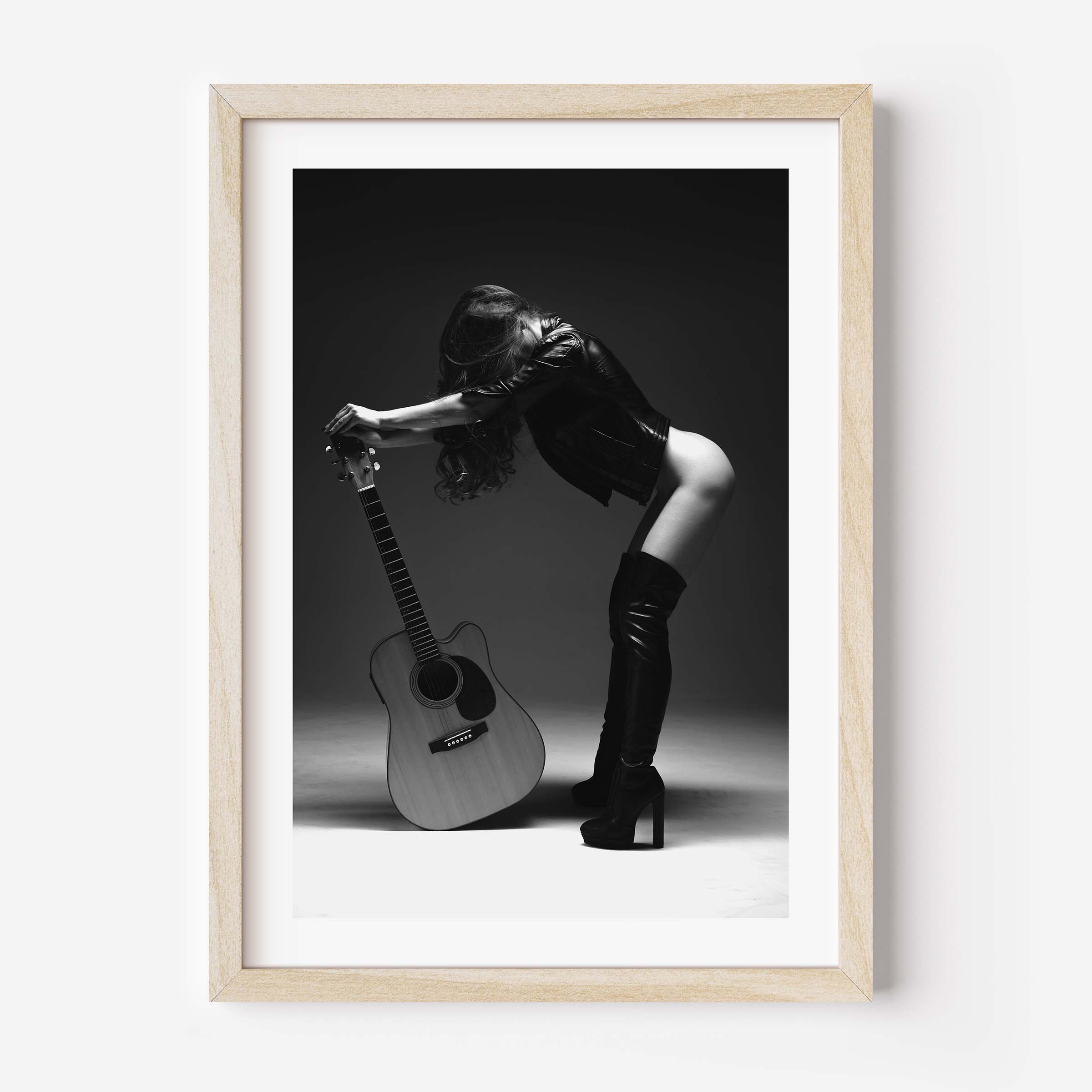 Black-and-white photo of a woman in a leather jacket and thigh-high boots leaning over an acoustic guitar in a studio, framed with white mat.