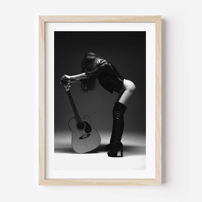 Black-and-white photo of a woman in a leather jacket and thigh-high boots leaning over an acoustic guitar in a studio, framed with white mat.