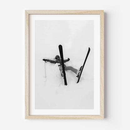 Black and white fine art print of a skier lying in deep snow with skis and pole scattered, framed in black.