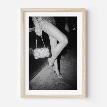 Black-and-white fine art photograph of a woman’s legs in high heels and sequin dress, holding a glittering handbag on a city sidewalk.