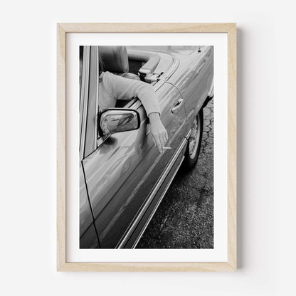 A framed black and white art print depicting a person in a convertible car with their arm out the window, held by a light natural wood frame and white matting, shown against a plain white wall.