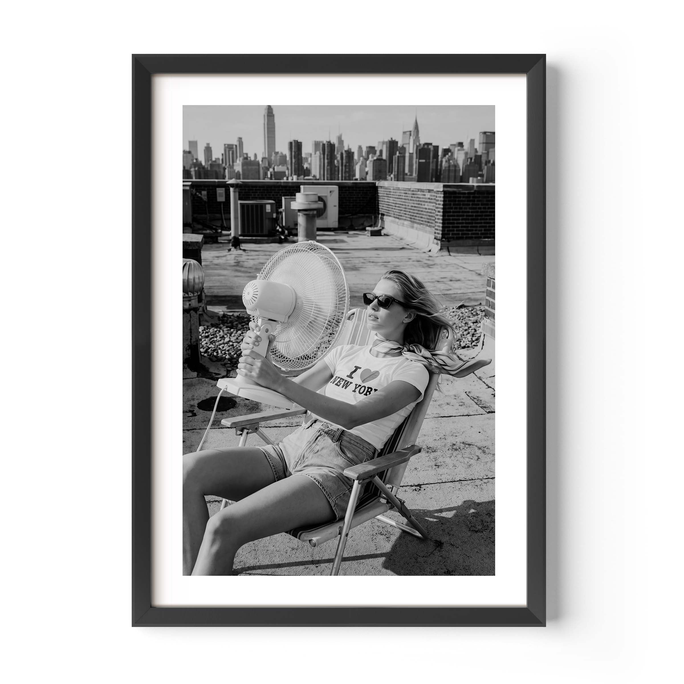 A framed black and white art print featuring a woman sitting on a folding chair on a rooftop with a city skyline in the background, displayed against a clean white wall.