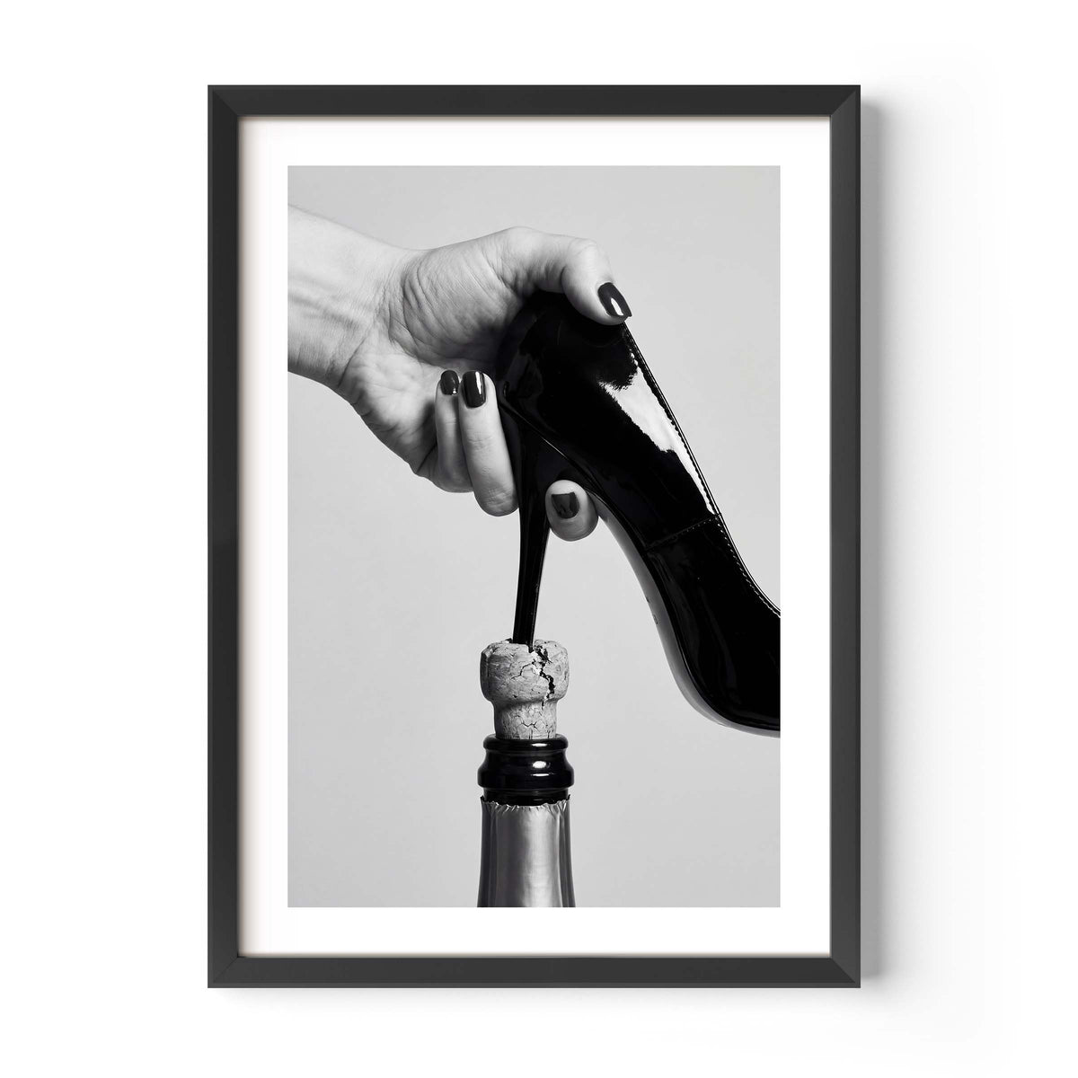 A framed black and white art print featuring a hand holding a high-heeled shoe above a champagne bottle, displayed on a clean white background.