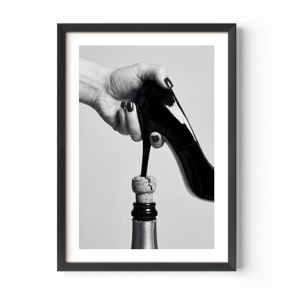 A framed black and white art print featuring a hand holding a high-heeled shoe above a champagne bottle, displayed on a clean white background.
