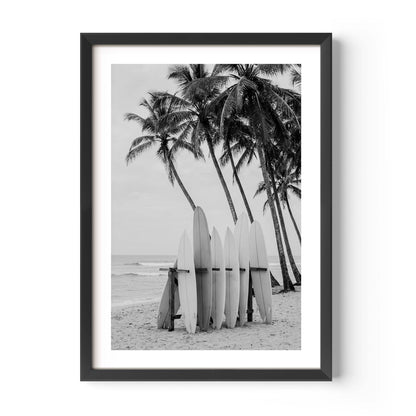 A framed black and white beach photograph featuring surfboards on the sand in front of palm trees, presented in a black frame with a white mat against a light gray background.
