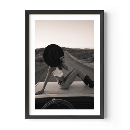 Fine art photo print of a woman sitting on a car roof on a desert road, holding a wide-brim hat in front of her face at sunset.