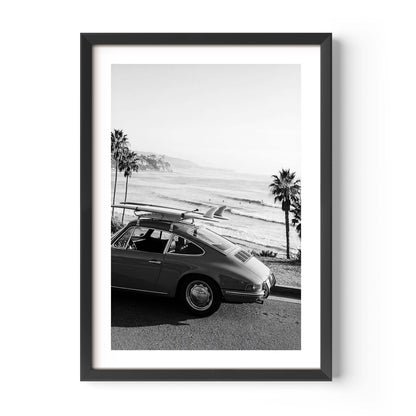 Black and white framed wall art print showing a classic porsche car with surfboards parked by the ocean with palm trees and coastline in the background.