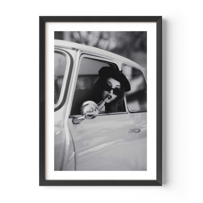 Black and white framed wall art print showing a woman in sunglasses sitting in a vintage car while applying lipstick.