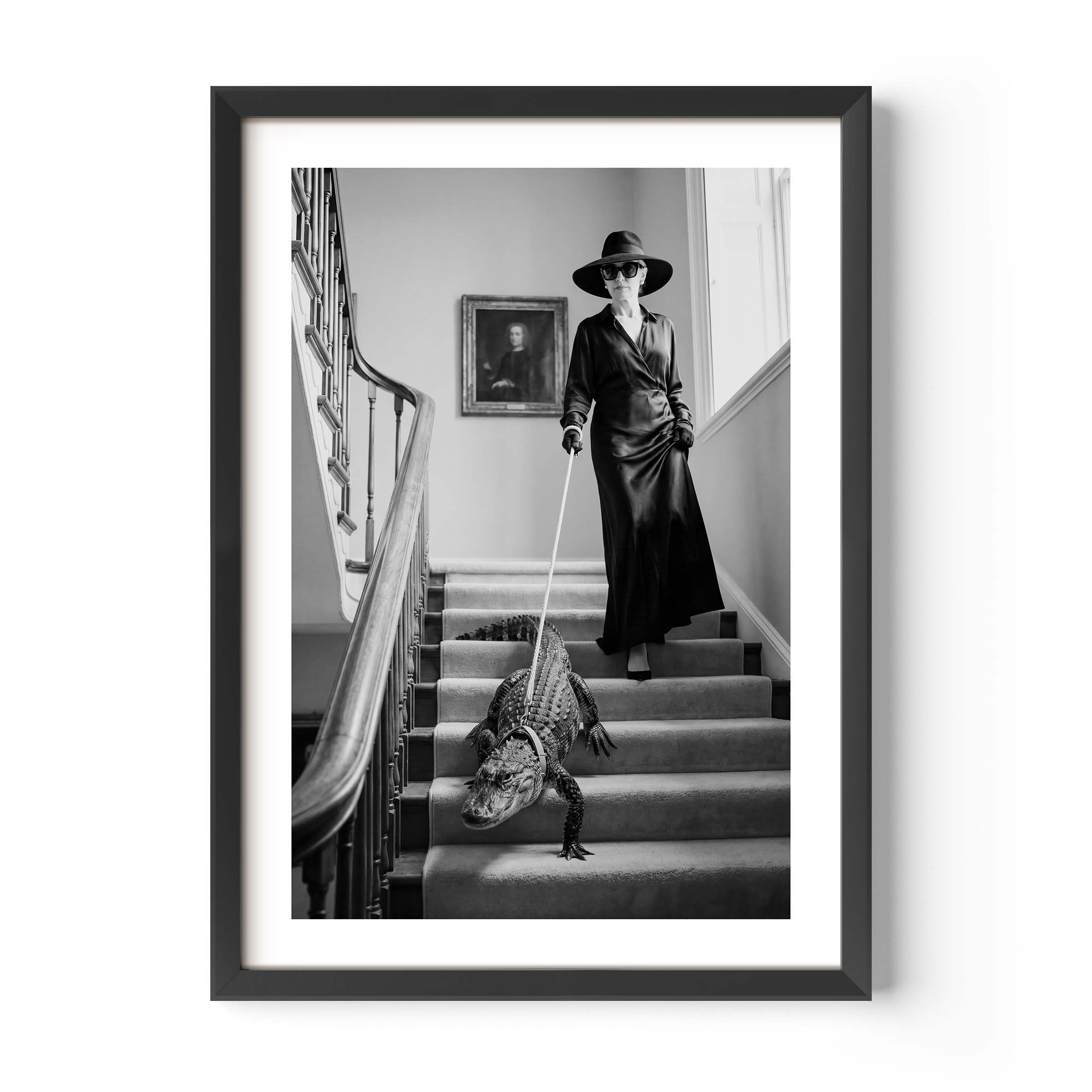A framed black and white art print depicting a woman in a long coat and hat walking an alligator up a staircase, shown against a clean white background.