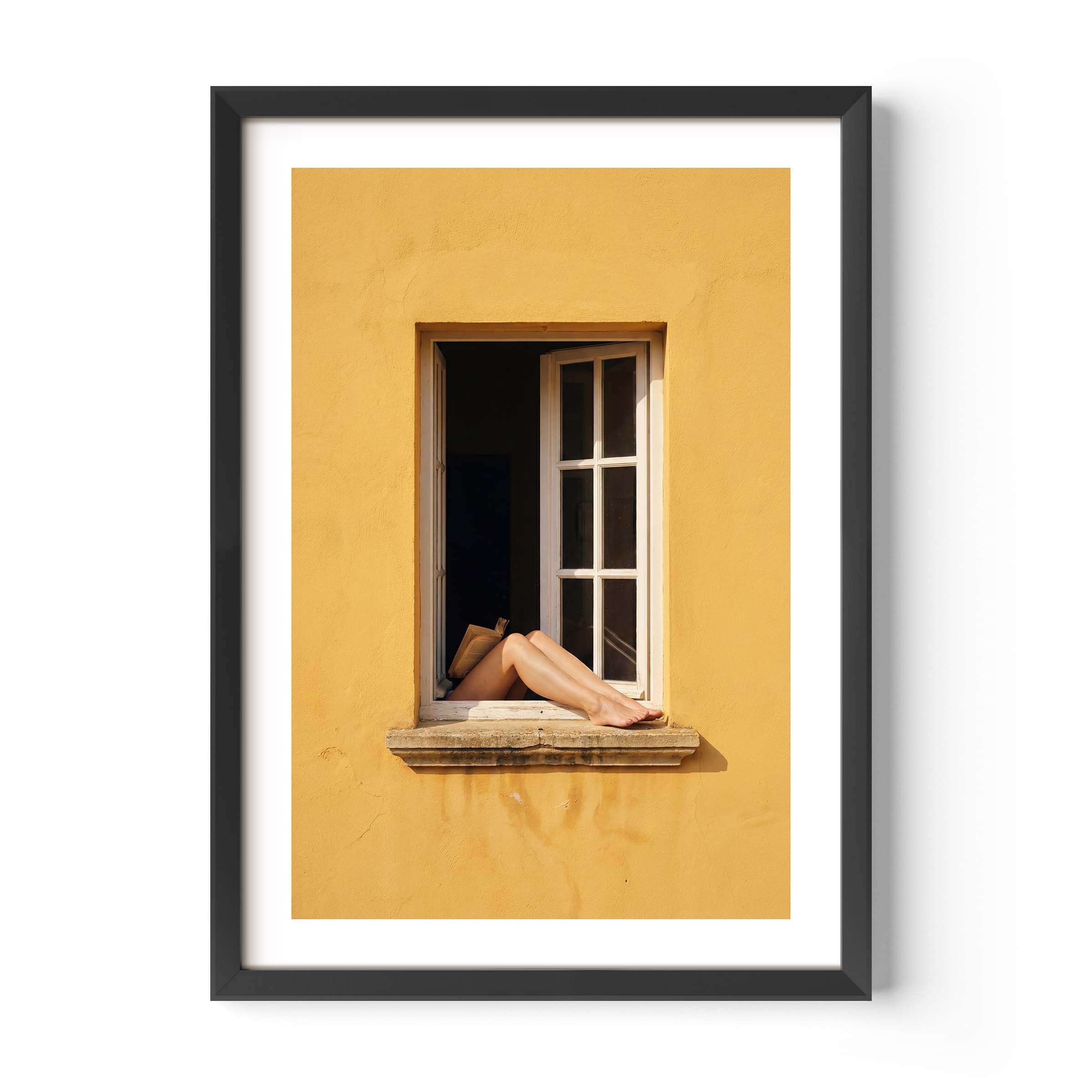 
Art print showing bare legs and feet resting on the stone sill of an open white-framed window set into a warm golden-yellow Mediterranean wall, in a natural wood frame with white mat border.