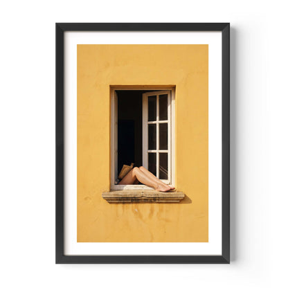 
Art print showing bare legs and feet resting on the stone sill of an open white-framed window set into a warm golden-yellow Mediterranean wall, in a natural wood frame with white mat border.
