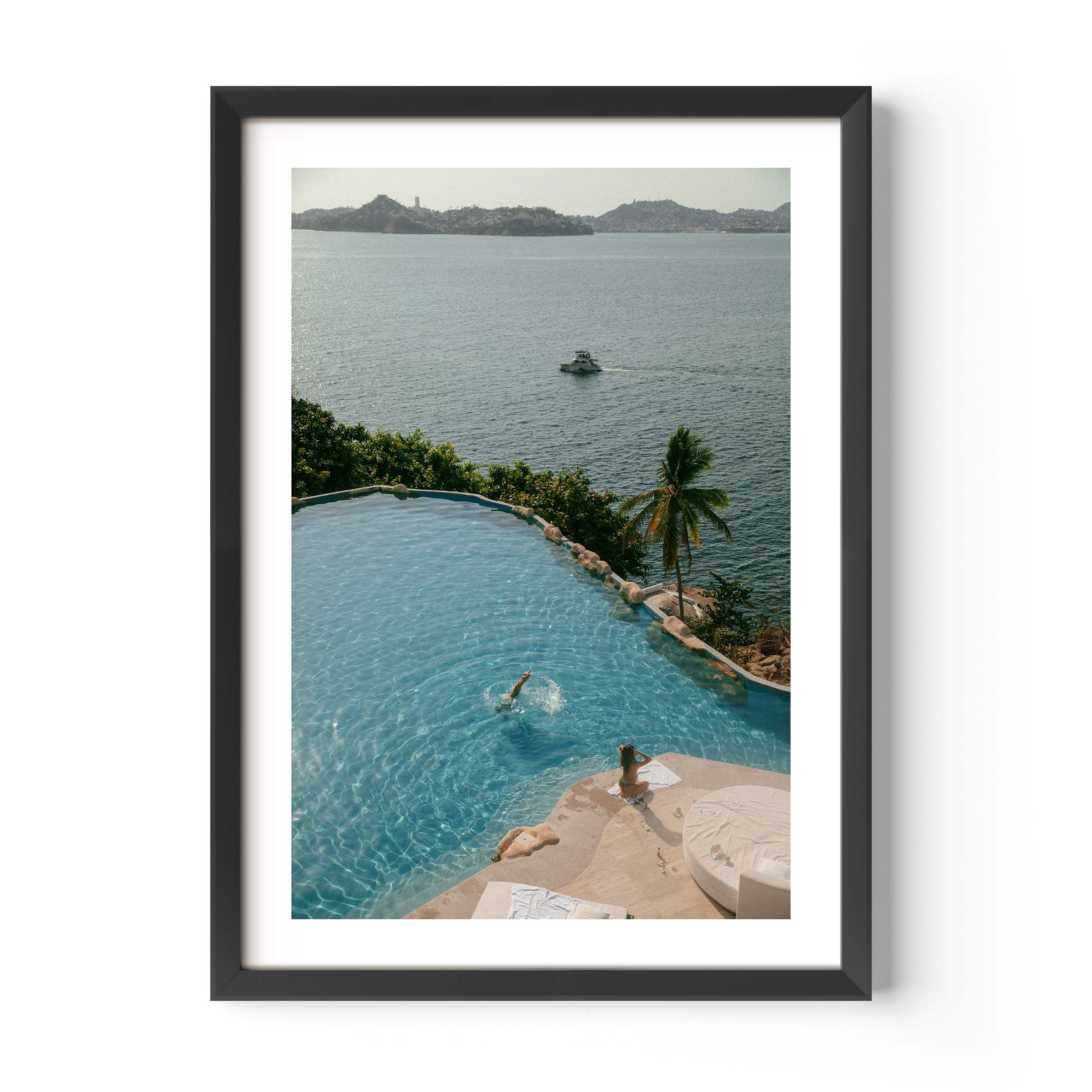 Framed color photograph of a diver in a turquoise infinity pool above a tropical bay with palm tree, sunlounger and boat in the distance — by Britt Van der Meyden.