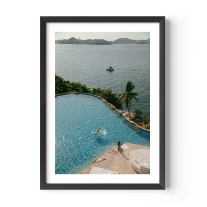 Framed color photograph of a diver in a turquoise infinity pool above a tropical bay with palm tree, sunlounger and boat in the distance — by Britt Van der Meyden.