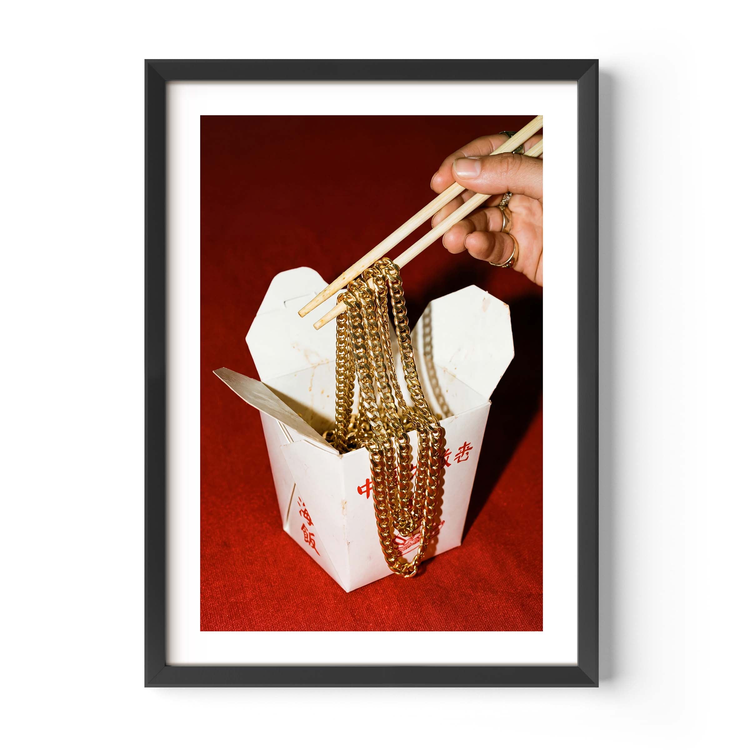 Conceptual photography print of a hand lifting gold chains with chopsticks from a Chinese takeout box on a red background, framed in black