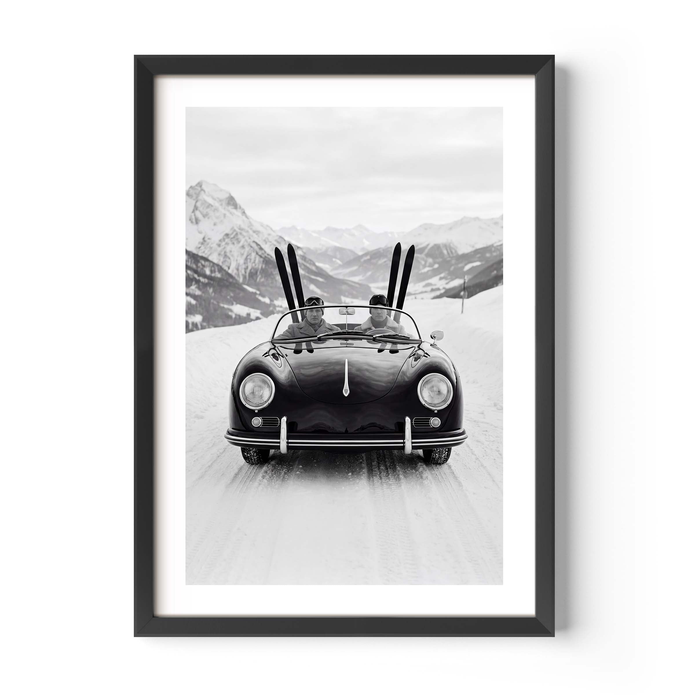 Black-and-white photo of a vintage convertible driving on a snowy mountain road, with two people inside and skis standing upright behind them, alpine peaks in the background.
