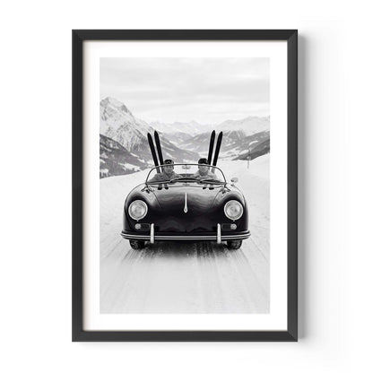 Black-and-white photo of a vintage convertible driving on a snowy mountain road, with two people inside and skis standing upright behind them, alpine peaks in the background.