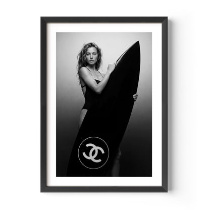 Black-and-white fine art photo of a woman in a swimsuit holding a large surfboard featuring a circular double-C style logo, against a minimal studio background.