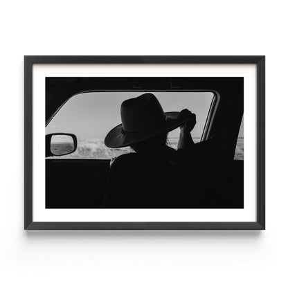 Black-and-white fine art print of a person wearing a cowboy hat seen from inside a car, looking out over an open landscape.