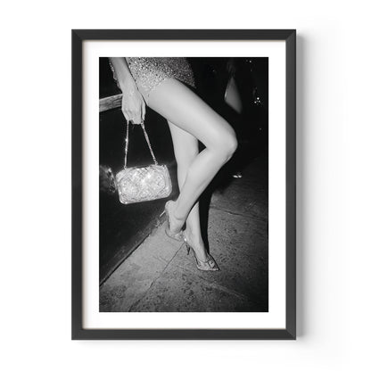 Black-and-white fine art photograph of a woman’s legs in high heels and sequin dress, holding a glittering handbag on a city sidewalk.