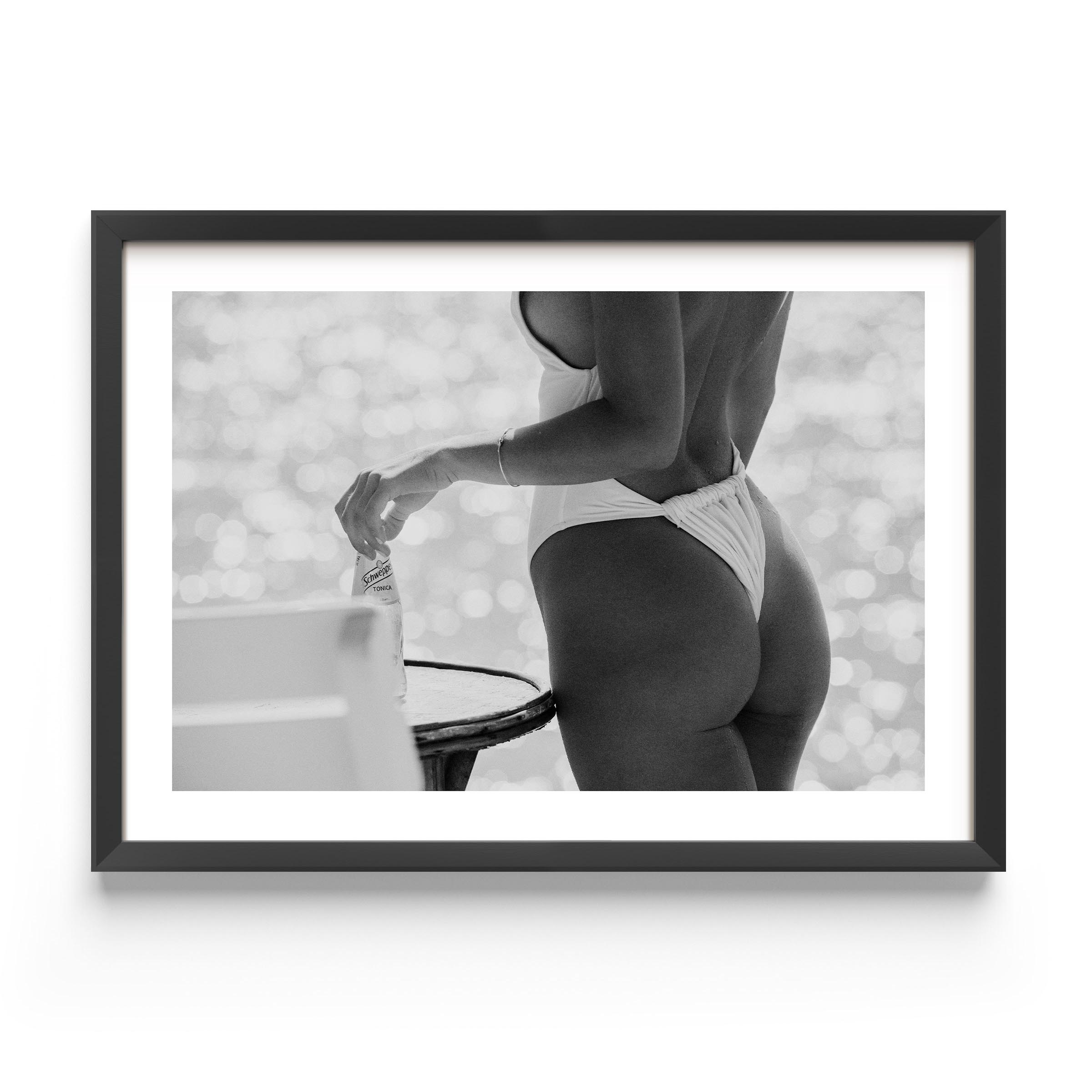 Black-and-white photo of a woman in a white one-piece holding a glass bottle by a sunlit seaside table