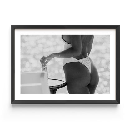 Black-and-white photo of a woman in a white one-piece holding a glass bottle by a sunlit seaside table