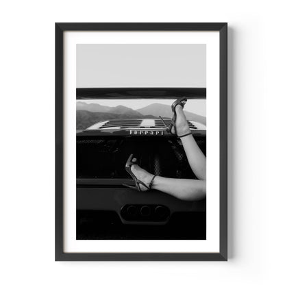 Black-and-white fine art print of high heels resting on the back of a Ferrari, framed wall art.