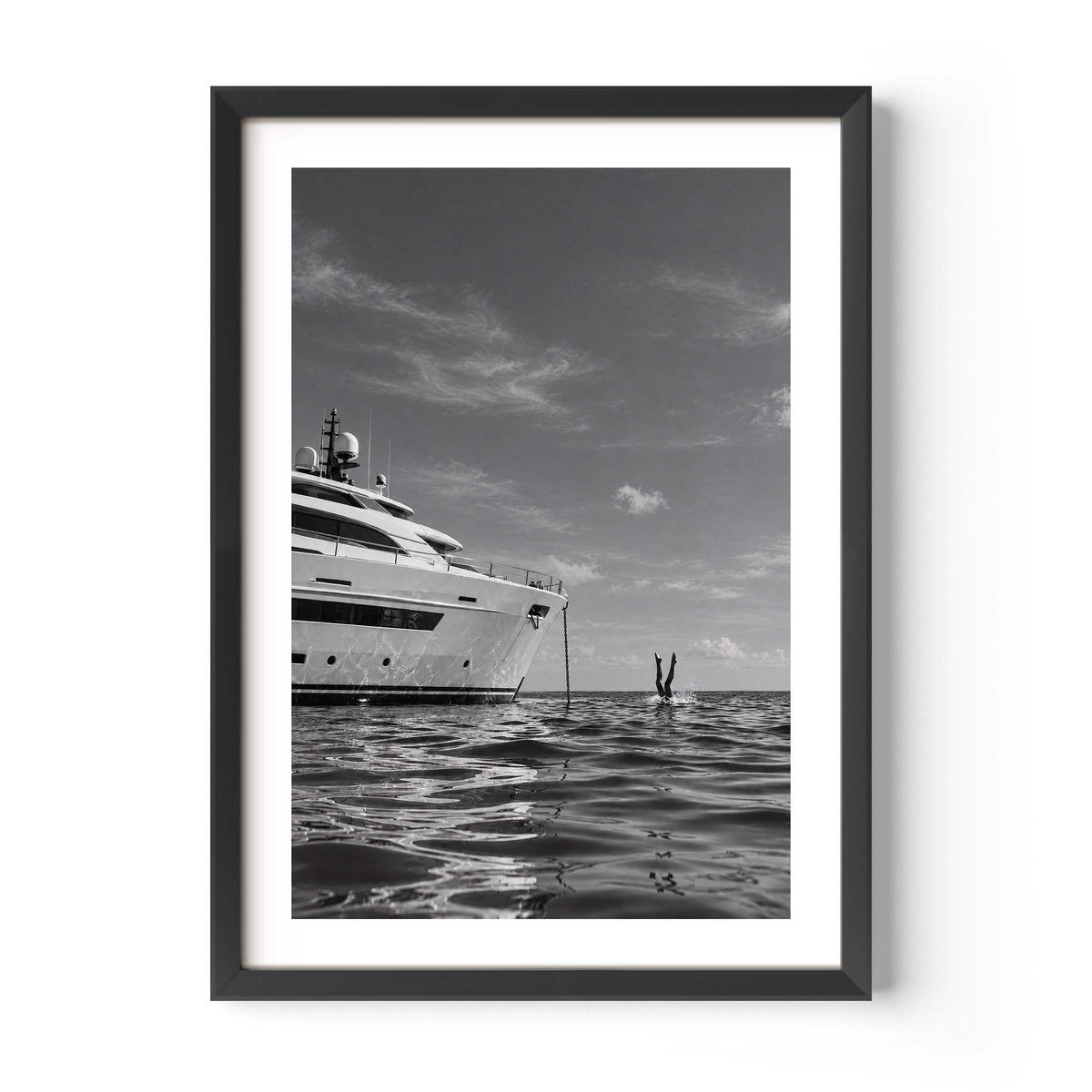 A framed black and white art print depicting a luxury yacht on calm water under a cloudy sky, with a small figure in the distance. The print is presented in a white mat and a modern black frame against a clean white background.