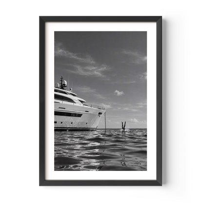 A framed black and white art print depicting a luxury yacht on calm water under a cloudy sky, with a small figure in the distance. The print is presented in a white mat and a modern black frame against a clean white background.